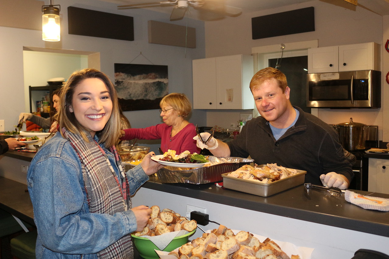 Photo of two people smiling at the camera and serving food. There are other people in the background not looking at the camera also serving food.