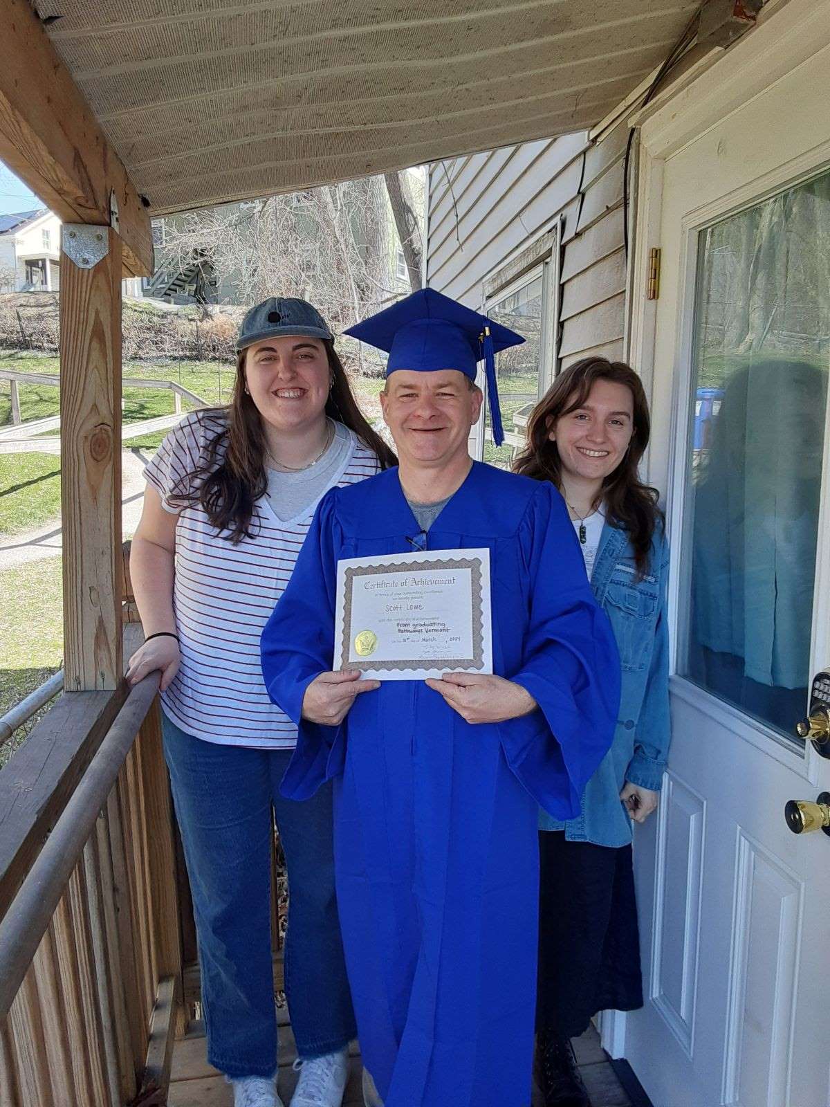 Photo of three people smiling at the camera. The person in the middle is wearing a blue cap and gown and celebration a graduation while holding a certificate. The person on the left is wearing a hat and a striped shirt. The person on the right is wearing a denim jacket.