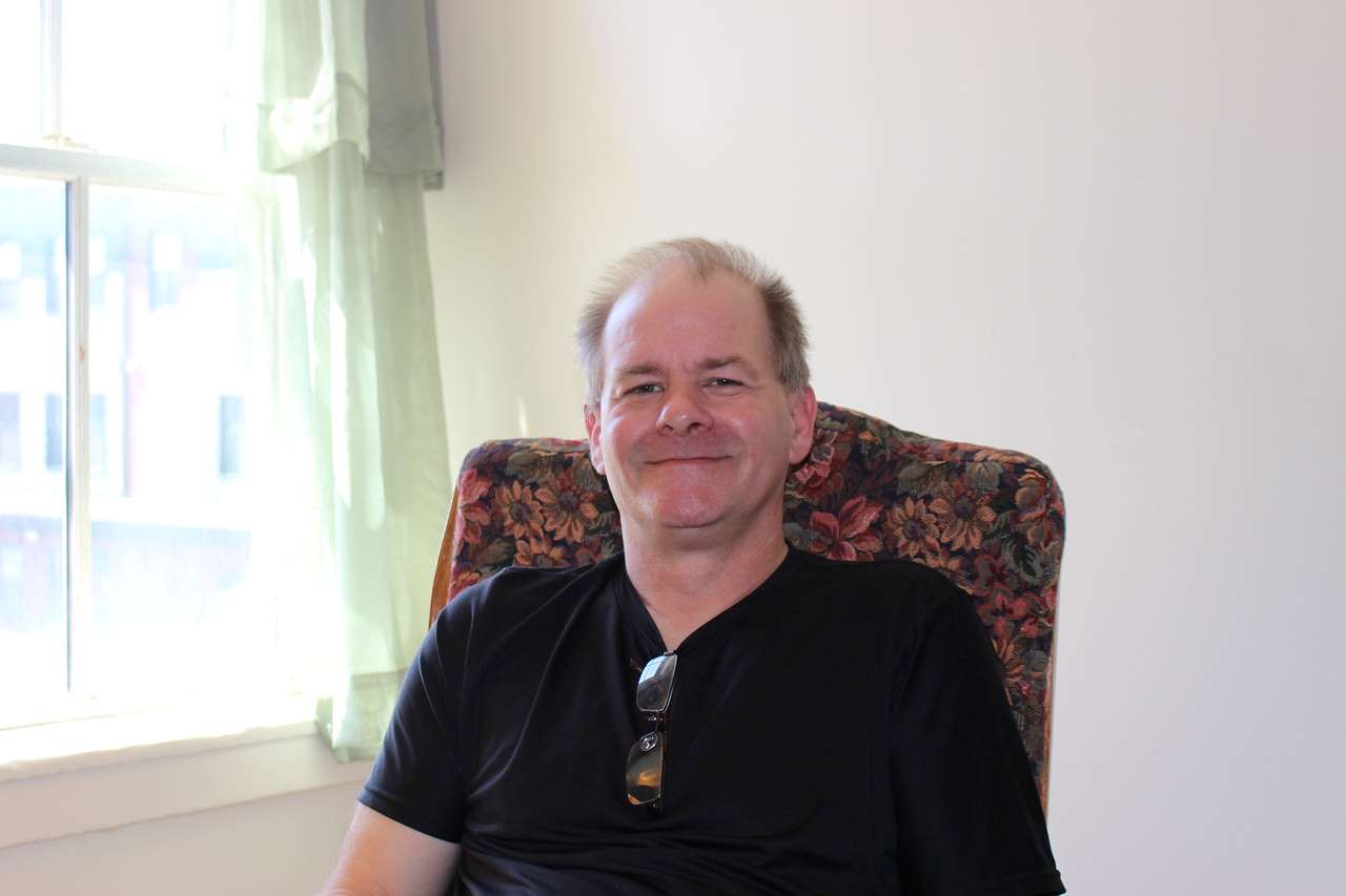 A man sitting in a red chair wearing a black shirt. He is sitting next to a window and is smiling.