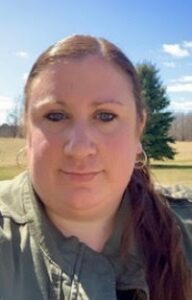 Photo of a woman with long brown hair outdoors on a sunny day, wearing a green jacket, with a field and trees in the background.