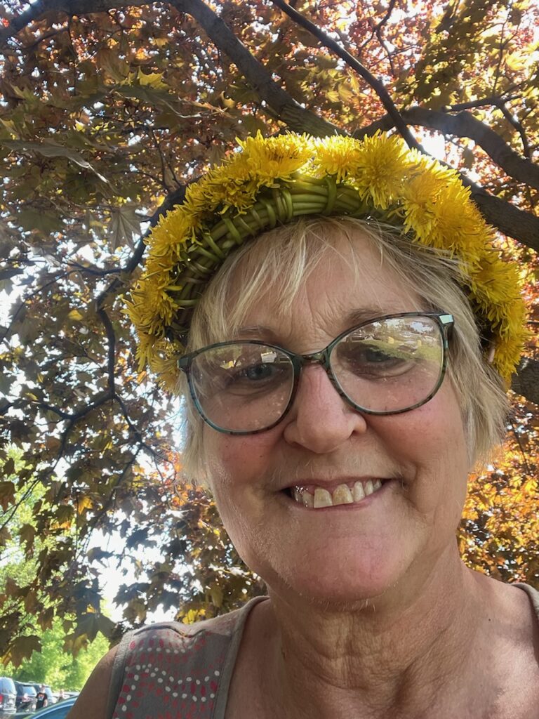 A smiling woman wearing glasses and a dandelion crown, standing under a tree with sunlight filtering through the leaves.