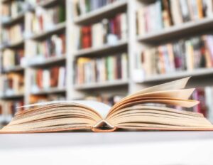 An open book on the table with shelves of books in the background.