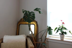 The image shows a cozy interior space with a beige armchair featuring a woven texture and a decorative pillow. Behind the chair, there is a gold-framed mirror resting against the wall. To the right, a large Monstera plant is positioned near the mirror, while a smaller potted plant with pink flowers sits on a windowsill, which is bathed in bright natural light. On a small table in front of the window, there is a tray holding several LED candles. The room has a calm and inviting atmosphere.