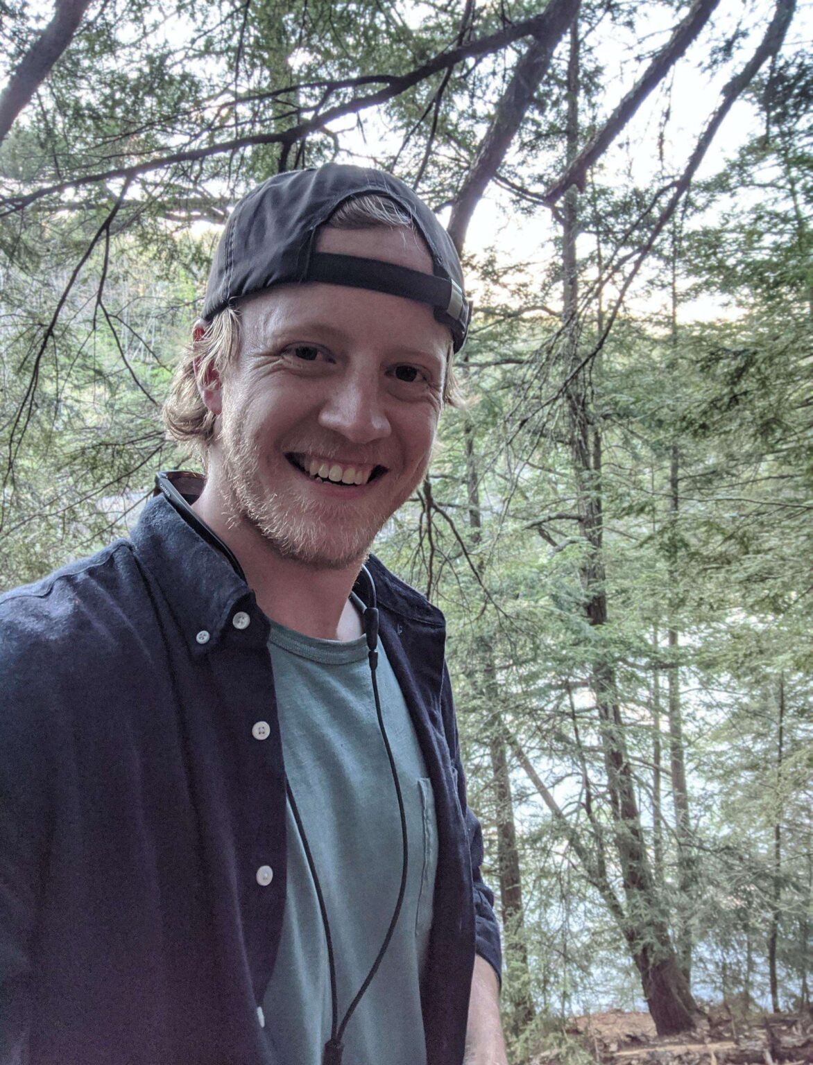 Selfie of a man with blonde hair wearing a baseball hat backwards. He is on a hike and is wearing a gray shirt with a darker gray jacket.