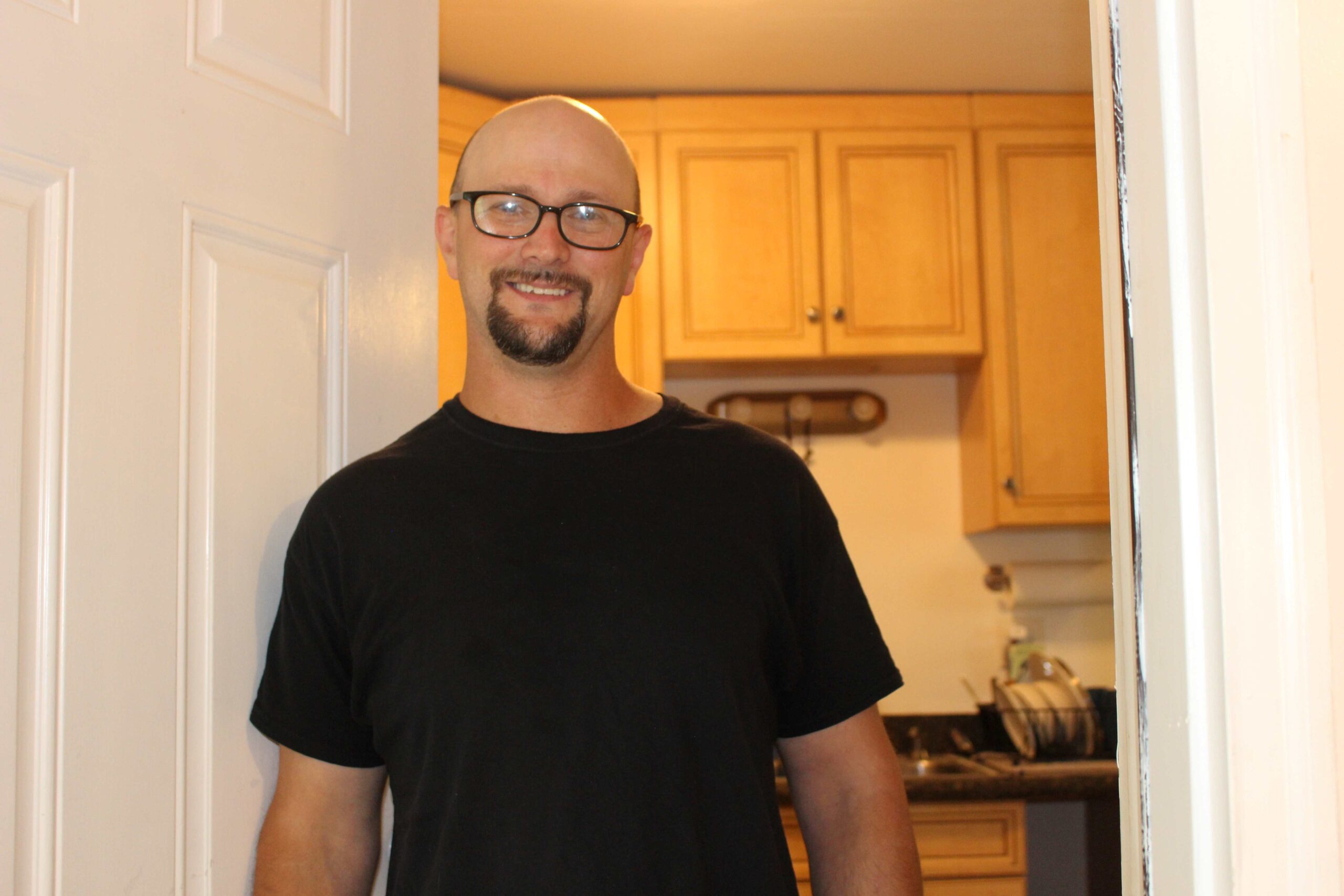 Photo of a man with glasses standing in the doorway to his apartment. He is wearing a black shirt.