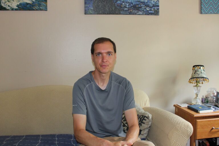 Photo of a man sitting on a couch wearing a gray t-shirt.